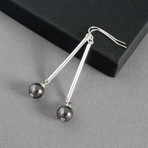 Puede incluir: Pendientes de plata con detalles de perlas negras. Los pendientes tienen una barra larga y delgada de plata con una perla negra en el extremo.
