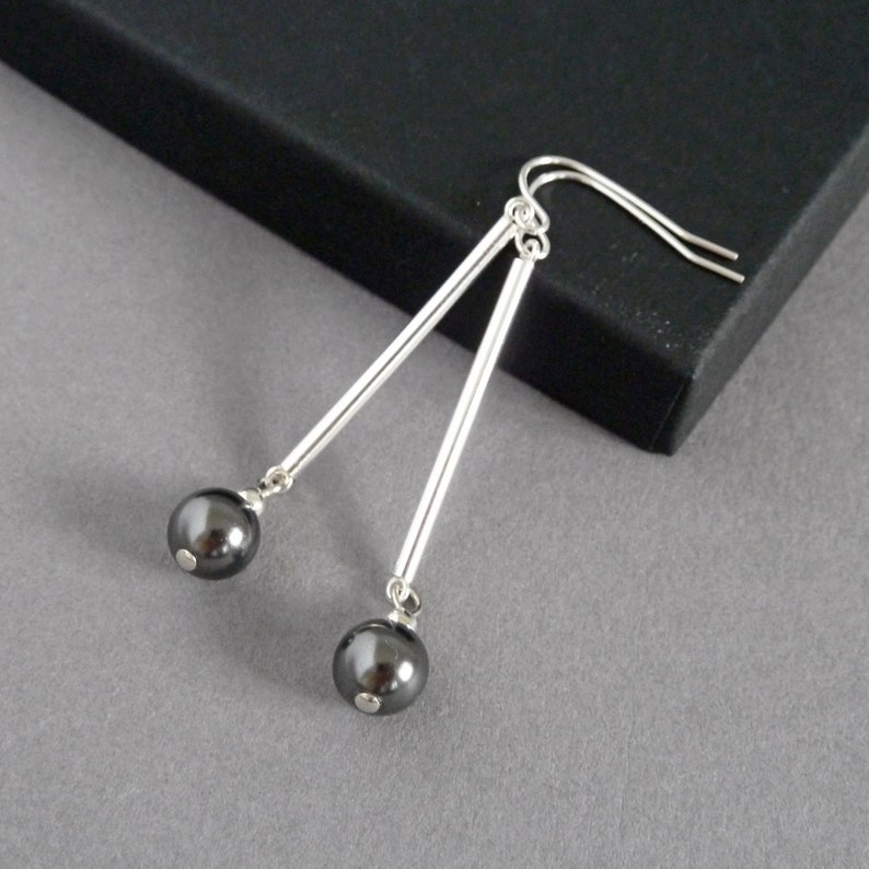 Puede incluir: Pendientes de plata con detalles de perlas negras. Los pendientes tienen una barra larga y delgada de plata con una perla negra en el extremo.