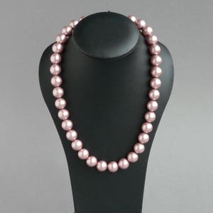 May include: A pink pearl necklace displayed on a black jewelry bust. The necklace features large, round, light pink pearls strung together. The pearls have a smooth, lustrous finish, and the necklace is draped in a gentle curve.