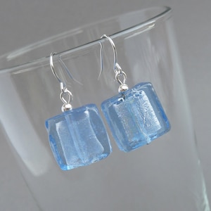 Può includere: Un paio di orecchini quadrati in vetro azzurro con ganci argentati. Gli orecchini sono di colore azzurro chiaro e sembrano essere fatti di vetro. Gli orecchini sono esposti davanti a un bicchiere trasparente.