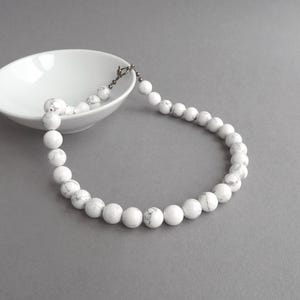 Puede incluir: Collar de cuentas blancas con cierre plateado, exhibido cerca de un cuenco blanco sobre una superficie gris. El collar presenta grandes cuentas blancas redondas con sutiles vetas grises, creando un diseño simple pero elegante.
