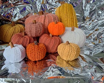 Pode incluir: Uma coleção de abóboras de crochê feitas à mão em vários tamanhos e cores, incluindo laranja, branco, amarelo e tons de rosa e marrom. Cada abóbora tem um pequeno caule marrom. As abóboras são dispostas em uma superfície prateada brilhante.