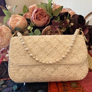 May include: Cream-colored handbag with a floral pattern and a pearl beaded handle. The bag has a flap closure and gold-colored hardware. A bouquet of pink and burgundy flowers is in the background.