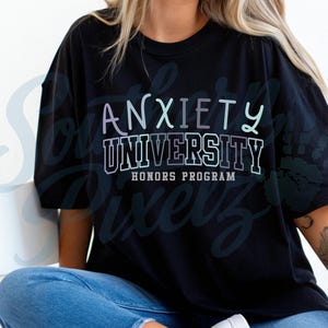 May include: Black t-shirt with the text "ANXIETY UNIVERSITY HONORS PROGRAM" in a layered, pastel font. The shirt is worn by a person in blue jeans.