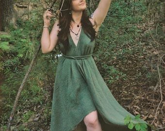 May include: A sleeveless, floor-length olive green dress with a deep V-neckline and a wrap-style waist tie. The fabric features a textured, crinkled appearance, and the skirt has an asymmetrical hemline that is shorter in the front.