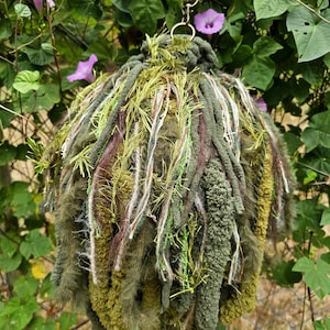 May include: A spherical hanging ornament crafted from a dense collection of green, olive, and brown yarn strands. The piece features varied textures including fuzzy, braided, and metallic fibers, suspended by a metal ring and a green fabric ribbon.