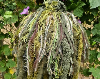 May include: A spherical hanging ornament crafted from a dense collection of green, olive, and brown yarn strands. The piece features varied textures including fuzzy, braided, and metallic fibers, suspended by a metal ring and a green fabric ribbon.