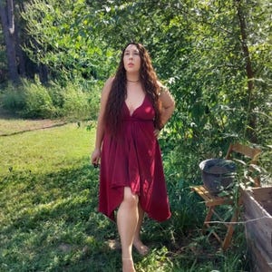 May include: A burgundy-colored dress with a plunging neckline and asymmetrical hemline. The dress is sleeveless and appears to be made of a flowing fabric. The image is taken outdoors, with a background of green foliage and grass.
