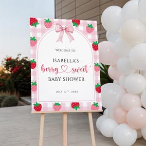 May include: A pink and white sign on a wooden easel announces "Welcome to Isabella's berry sweet BABY SHOWER." The sign features a pink gingham border, strawberry illustrations, and a pink bow. The date is July 12th, 20XX.