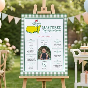 Könnte beinhalten: Ein grün-weiß kariertes Schild auf einer Holzstaffelei. Das Schild zeigt ein Babyfoto und Text, einschließlich der Worte "Mastered His First Year". Das Schild enthält auch die Spitznamen, Maße, das Lieblingsbuch, das Lied und das Essen des Babys.