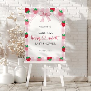 May include: A pink and white gingham baby shower sign on a white easel. The sign reads "Welcome to Isabella's berry sweet baby shower" with strawberry illustrations and a pink bow.