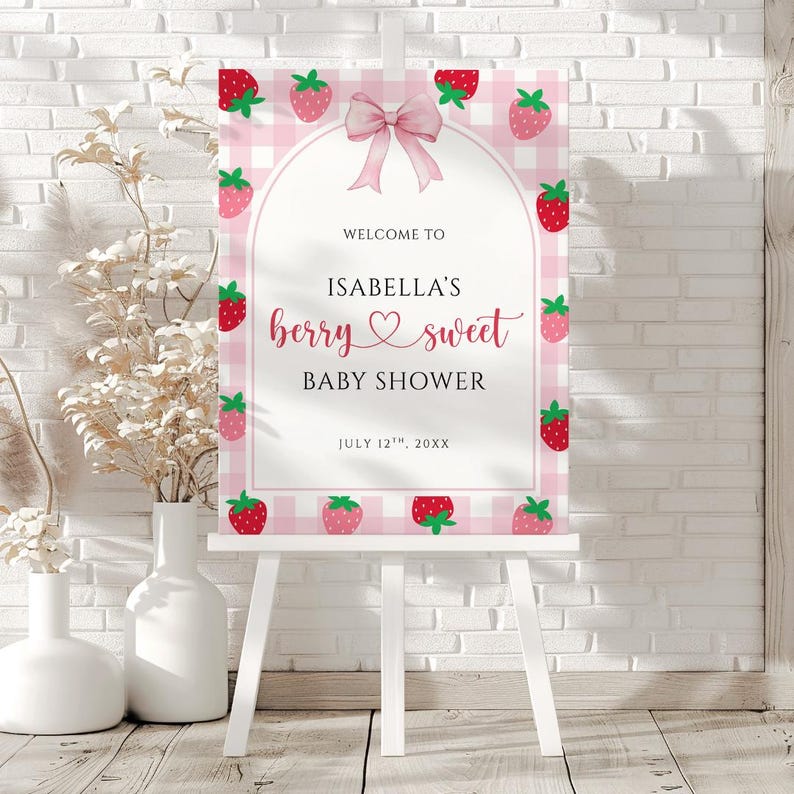 May include: A pink and white gingham baby shower sign on a white easel. The sign reads "Welcome to Isabella's berry sweet baby shower" with strawberry illustrations and a pink bow.