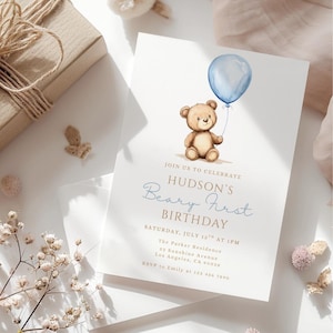 May include: A light-colored invitation card with a watercolor illustration of a teddy bear holding a blue balloon. The text reads "Hudson's Beary First Birthday" with event details. A gift box and floral accents are in the background.