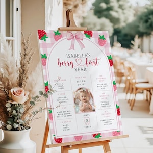 May include: A pink and white strawberry-themed first birthday sign on an easel. The sign features the text "Isabella's berry first year" with a photo and details about the child, including weight, height, and favorite things.