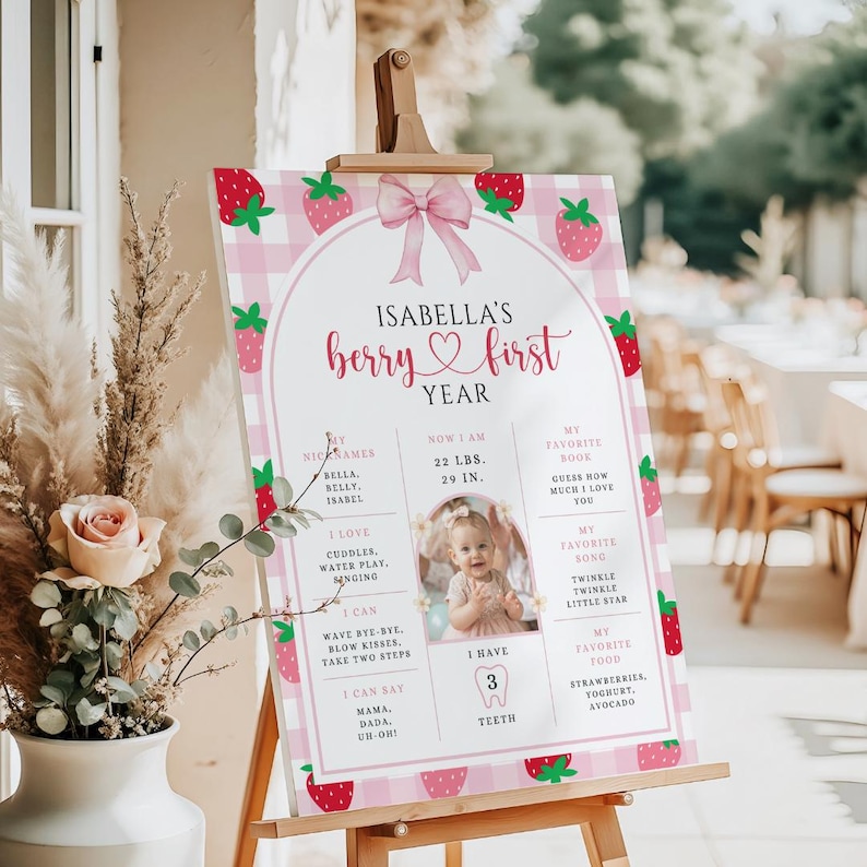 May include: A pink and white strawberry-themed first birthday sign on an easel. The sign features the text "Isabella's berry first year" with a photo and details about the child, including weight, height, and favorite things.