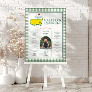 May include: A personalized milestone sign on a white easel with a green and white checkered border. The sign features a baby's photo, weight (22 lbs), height (29 in), and other details. Text includes "MASTERED His First Year" and "Hudson".