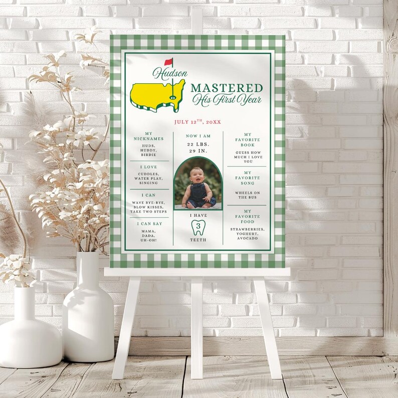 May include: A personalized milestone sign on a white easel with a green and white checkered border. The sign features a baby's photo, weight (22 lbs), height (29 in), and other details. Text includes "MASTERED His First Year" and "Hudson".