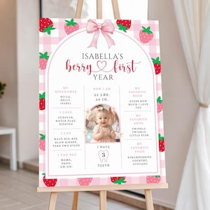 May include: A pink and white strawberry-themed first birthday sign on an easel. The sign features the text "Isabella's berry first year" with a photo of a baby. The sign includes the baby's nicknames, favorite things, and measurements: 22 lbs, 29 in.