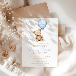 May include: A light beige invitation card with a watercolor illustration of a teddy bear holding a blue balloon. The text reads "Join us to celebrate Hudson's Beary First Birthday" with event details. The card is on a neutral background.