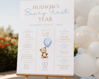 May include: A white sign on an easel with the text "Hudson's Beary First Year." The sign includes a teddy bear graphic, a blue balloon, and details about the child's first year, including weight, height, and favorite things.