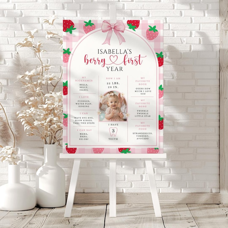 May include: A pink and white strawberry-themed first year milestone poster on a white easel. The poster features the text "Isabella's berry first year" and includes a photo, weight, height, and favorite things. The poster is decorated with strawberry illustrations.