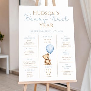 May include: A white sign on an easel with the text "Hudson's Beary First Year." The sign includes a teddy bear holding a blue balloon, and details such as nicknames, favorite book, and favorite food. The sign states the child is 22 lbs and 29 inches.
