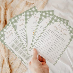 May include: A stack of baby shower game cards with a green and white gingham border. The cards feature various games, including a word scramble and trivia questions. The text on the cards is in a handwritten style.