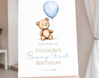 May include: A white sign on an easel features a watercolor illustration of a teddy bear holding a blue balloon. Text reads "Welcome to Hudson's Beary First Birthday" with a date below.