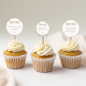 May include: Three cupcakes with white frosting and decorative toppers. The toppers read "Welcome Baby Parker", "Isabella's Baby Shower", and "Locally Grown Baby On The Way". The cupcakes are in white paper liners.
