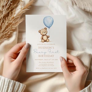 May include: A light-colored invitation card with a watercolor illustration of a teddy bear holding a blue balloon. The text reads "Hudson's Beary First Birthday" with event details. The card is held by a person.
