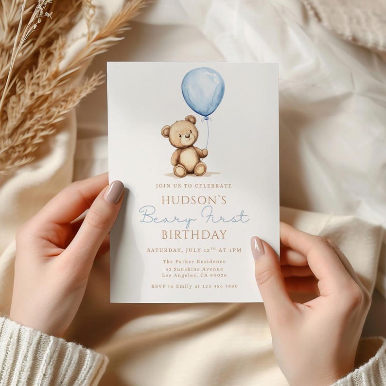 May include: A light-colored invitation card with a watercolor illustration of a teddy bear holding a blue balloon. The text reads "Hudson's Beary First Birthday" with event details. The card is held by a person.