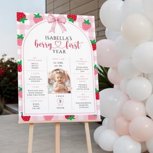 May include: A pink and white sign on a wooden easel, decorated with strawberries and a pink bow. The sign reads "Isabella's berry first year" and includes details about the child's nicknames, weight, height, favorite book, song, and food.