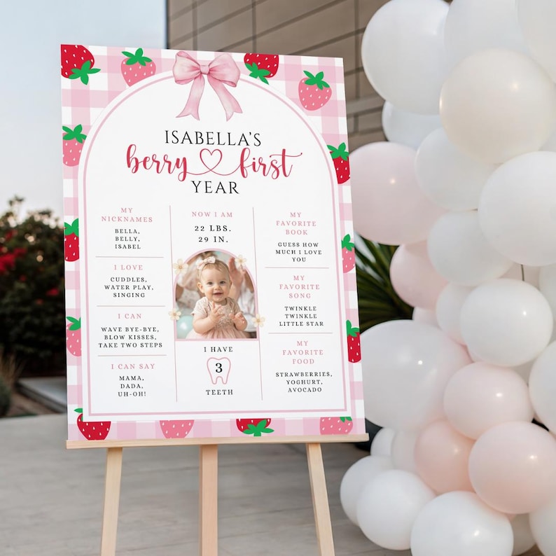 May include: A pink and white sign on a wooden easel, decorated with strawberries and a pink bow. The sign reads "Isabella's berry first year" and includes details about the child's nicknames, weight, height, favorite book, song, and food.