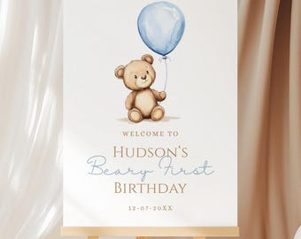 May include: A white sign on an easel featuring a watercolour illustration of a teddy bear with a blue balloon. The sign says "Welcome to Hudson's Beary First Birthday" in blue script, with a date below.