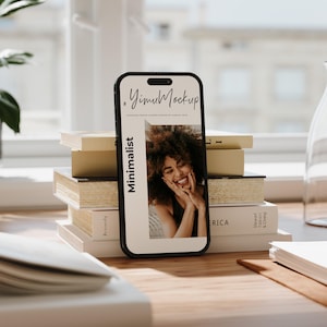 May include: A black smartphone displays a mockup with the text "YimuMockup" and "Minimalist." The screen shows a smiling woman with curly hair. The phone rests on a wooden surface next to a stack of books.