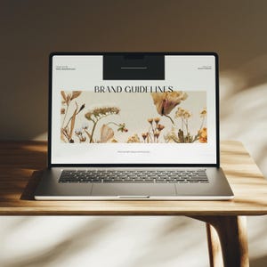 Peut inclure: Un ordinateur portable argenté affichant une page web avec le texte "BRAND GUIDELINES" et des illustrations florales. L'ordinateur est posé sur une table en bois, avec un effet de lumière et d'ombre. L'écran affiche également le texte "Premium Professional Mockups".