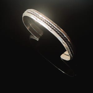 Könnte beinhalten: Ein silberfarbenes Armband in Manschettenform mit einem mittig verlaufenden, gedrehten Seilmuster entlang der gesamten Schiene. Die Metalloberfläche ist poliert und weist feine, eingravierte Details entlang der Kanten des Seilmotivs auf.