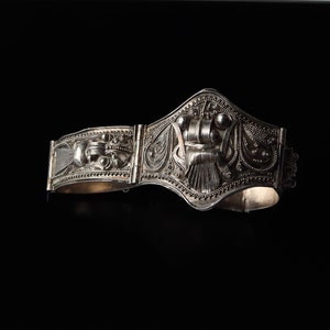 May include: A silver bracelet featuring intricate floral and geometric designs. The bracelet is hinged and has a decorative clasp.