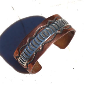 Op de afbeelding: Koperen manchetarmband met een centrale zilverkleurige band en gedetailleerde geëtste ontwerpen. De armband heeft een warme, roodbruine tint met ingewikkelde patronen langs de randen en in het midden, wat een opvallend contrast creëert.