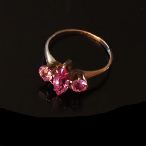 May include: A gold ring featuring a central marquise-cut pink gemstone flanked by two smaller round-cut pink gemstones. The band is thin and polished, showcasing a classic three-stone design with vibrant pink stones set in a gold metal setting.