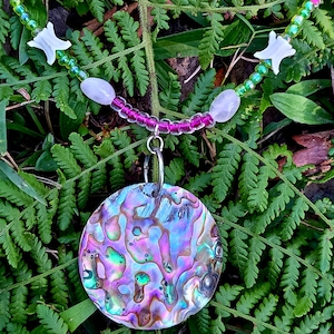 May include: Iridescent abalone shell pendant necklace. The round pendant hangs from a beaded strand with pink, green, and white beads. The necklace is displayed on a bed of green fern leaves.