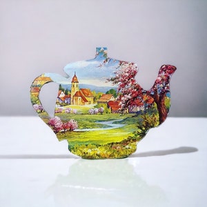 Pode incluir: Uma chaleira de cerâmica com uma cena pintada colorida de uma aldeia com uma igreja, árvores e flores. A chaleira é branca com um esmalte azul claro e tem uma alça e um bico.
