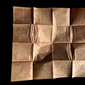 May include: A folded sheet of tan paper with a floral design in red ink. The paper is divided into sixteen sections, each featuring a different floral illustration. Some sections contain text. The paper is against a black background.