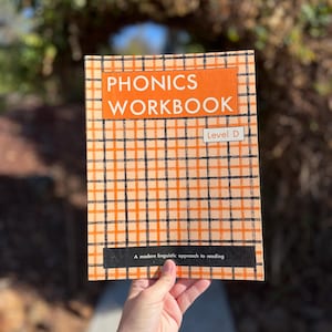 Peut inclure: Un cahier d'exercices de phonétique, Phonics Workbook Level D, avec un motif de grille composé de lignes orange et noires. La couverture affiche le texte PHONICS WORKBOOK en blanc sur fond orange et A modern linguistic approach to reading.