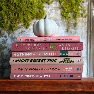 Puede incluir: Una pila de seis libros de bolsillo rosas con títulos de texto blanco. Los libros están apilados sobre una superficie de madera. Una calabaza blanca se sienta en la parte superior de la pila de libros.