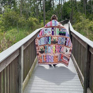 May include: A colorful handmade quilt with a bookshelf design is draped over a person walking on a wooden boardwalk. The quilt features various fabric patterns, including floral, striped, and solid colors. The background shows a natural setting with trees and greenery.