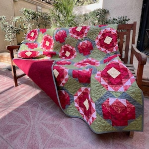 May include: A quilt featuring a repeating geometric pattern of red and pink floral-like shapes on a sage green background with a cream-colored center square in each motif. The quilt has a solid deep red underside and intricate swirl stitching.