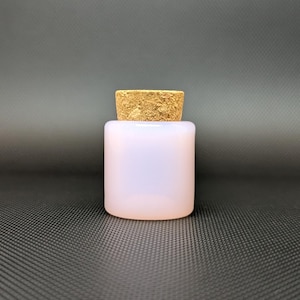 Pode incluir: Uma pequena garrafa cilíndrica de vidro fosco com uma rolha de cortiça. A garrafa é opaca e tem uma superfície lisa. A cortiça é de cor castanho claro e tem um aspeto texturizado. A garrafa está sobre uma superfície cinza escuro.