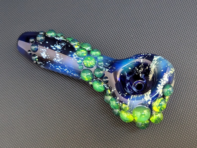 May include: A blue glass pipe with a galaxy design and green accents. The pipe is decorated with small, round, green glass beads.