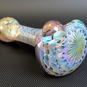 Puede incluir: Una pipa de vidrio soplado a mano con colores iridiscentes. La pipa presenta un cuenco con un patrón texturizado de formas de lágrima azules y verdes y un tallo con un diseño escamoso. Una burbuja clara y redondeada se encuentra encima del cuenco.